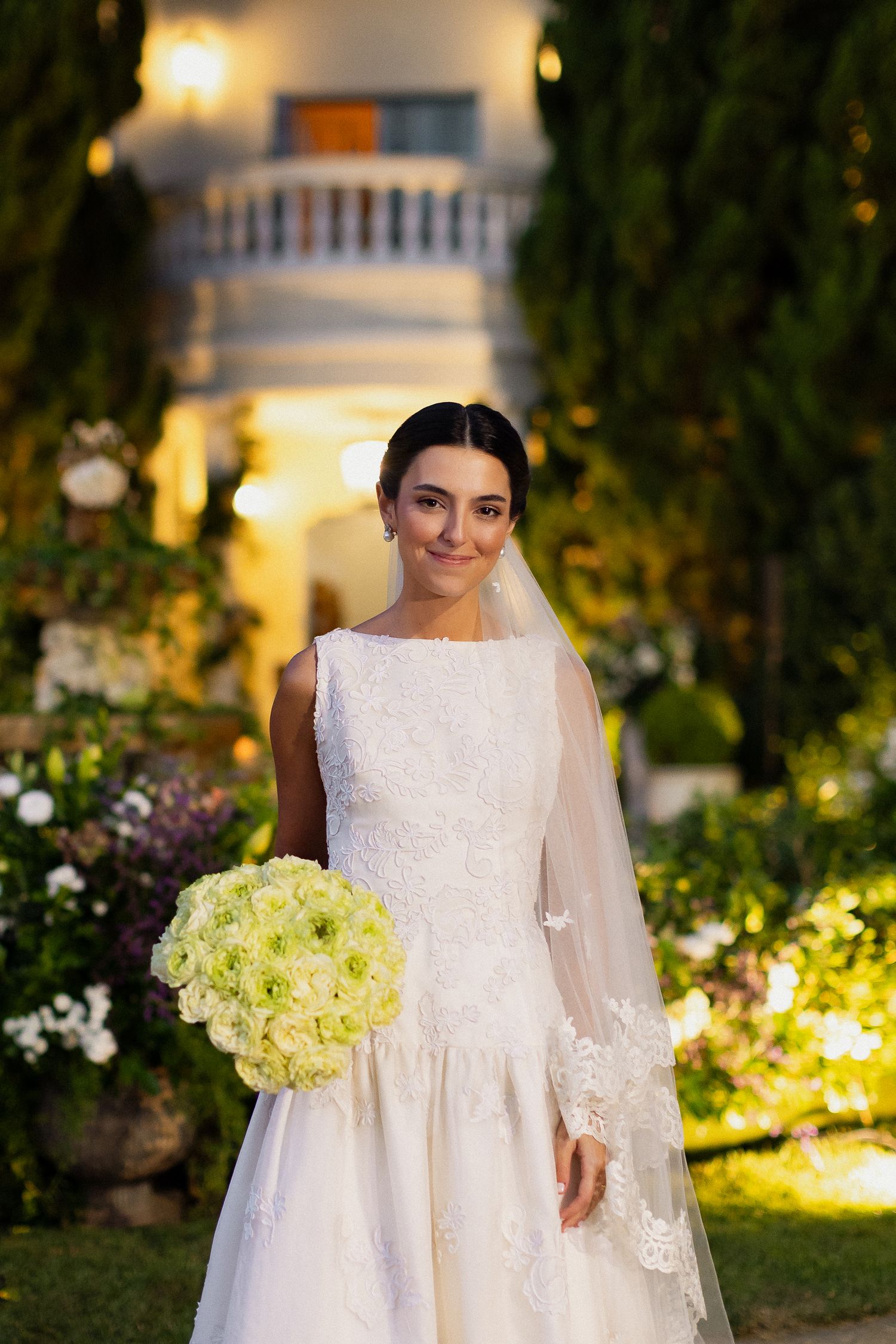 A bride in an elegant white drop-waist wedding dress holds a bouquet of cream roses in a garden at dusk.