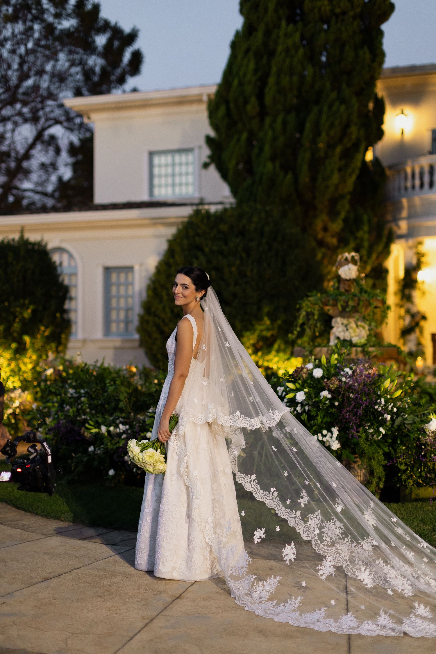 A bride in a white lace gown with a long embroidered cathedral veil stands in front of an elegant villa at dusk.