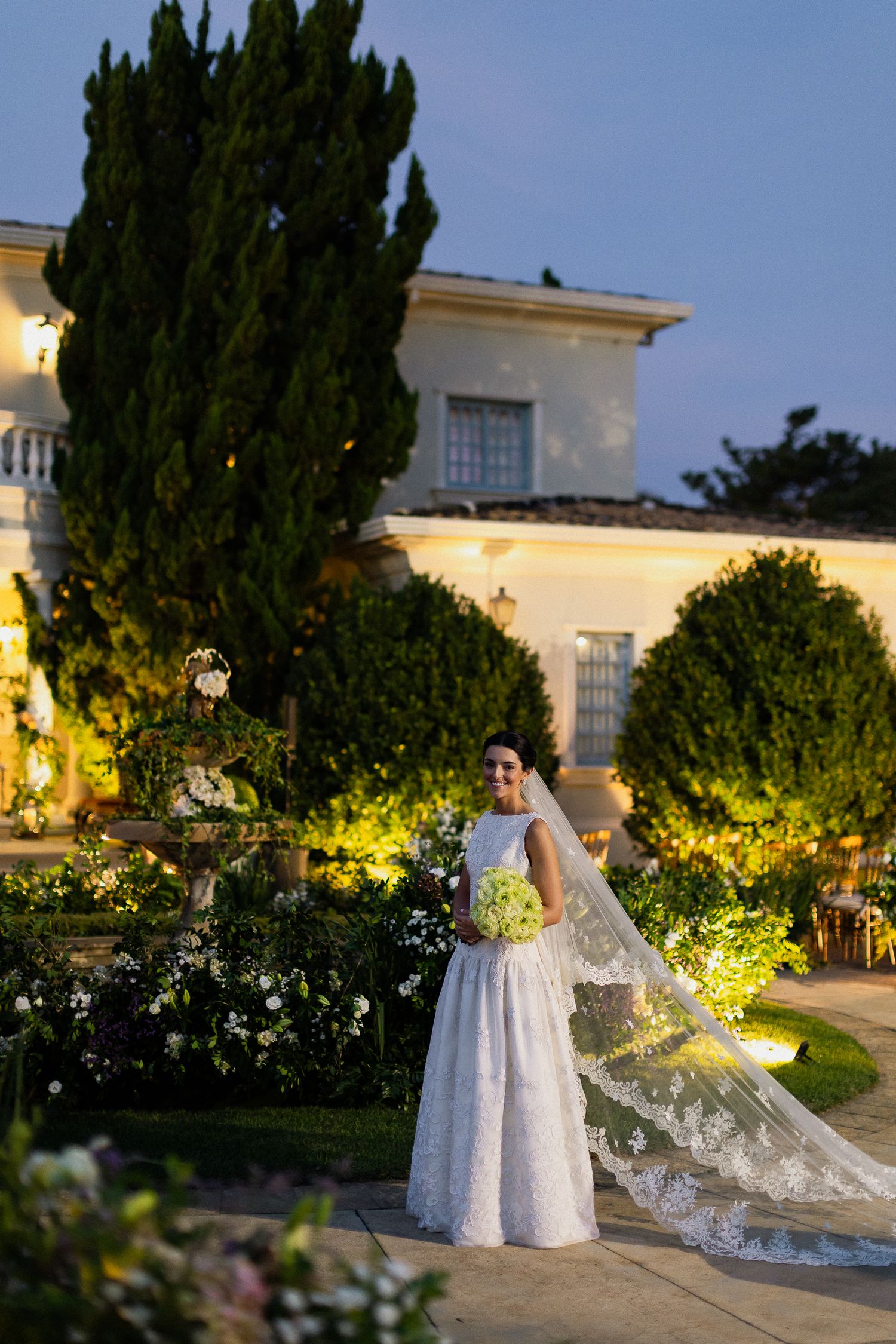 A bride in a flowing white gown poses in a garden setting at dusk with illuminated trees and architecture.