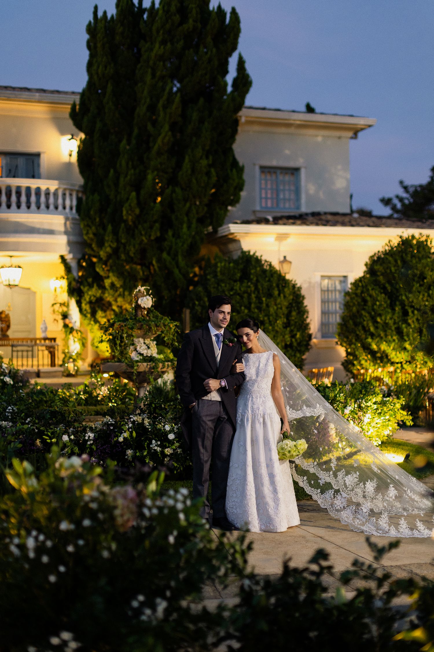 A couple poses in formal wedding attire at dusk in front of an elegant villa with illuminated gardens.