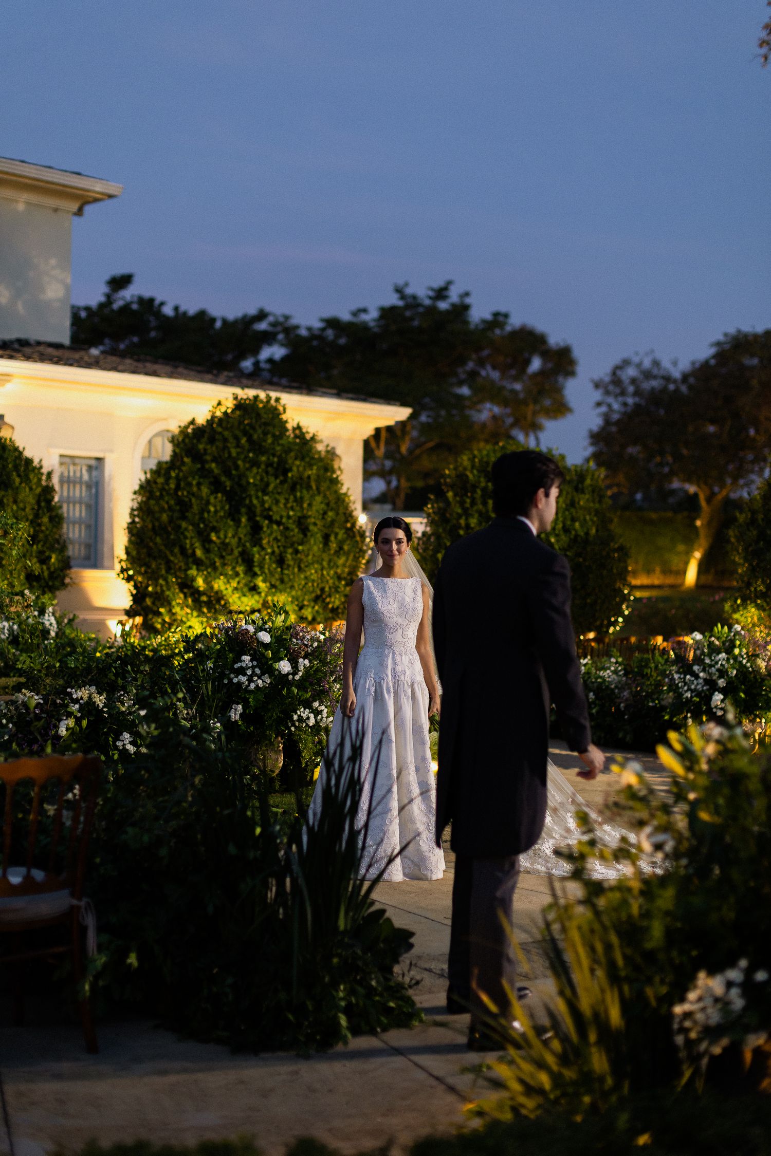 A couple in wedding attire walks along an illuminated garden path at dusk with lush greenery and flowers surrounding them.