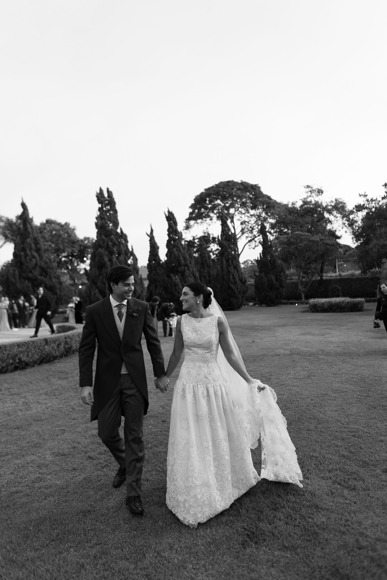 A bride and groom walk together on a garden lawn with tall cypress trees in the background in black and white.
