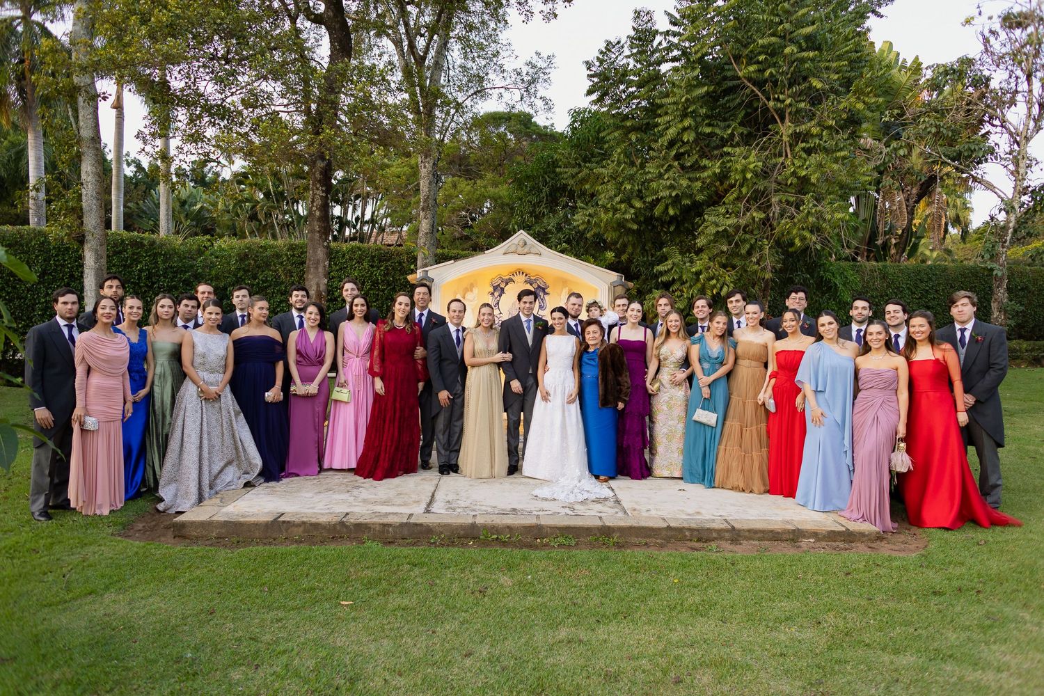 Large formal group photo at an outdoor wedding with attendees wearing colorful evening gowns and formal attire.