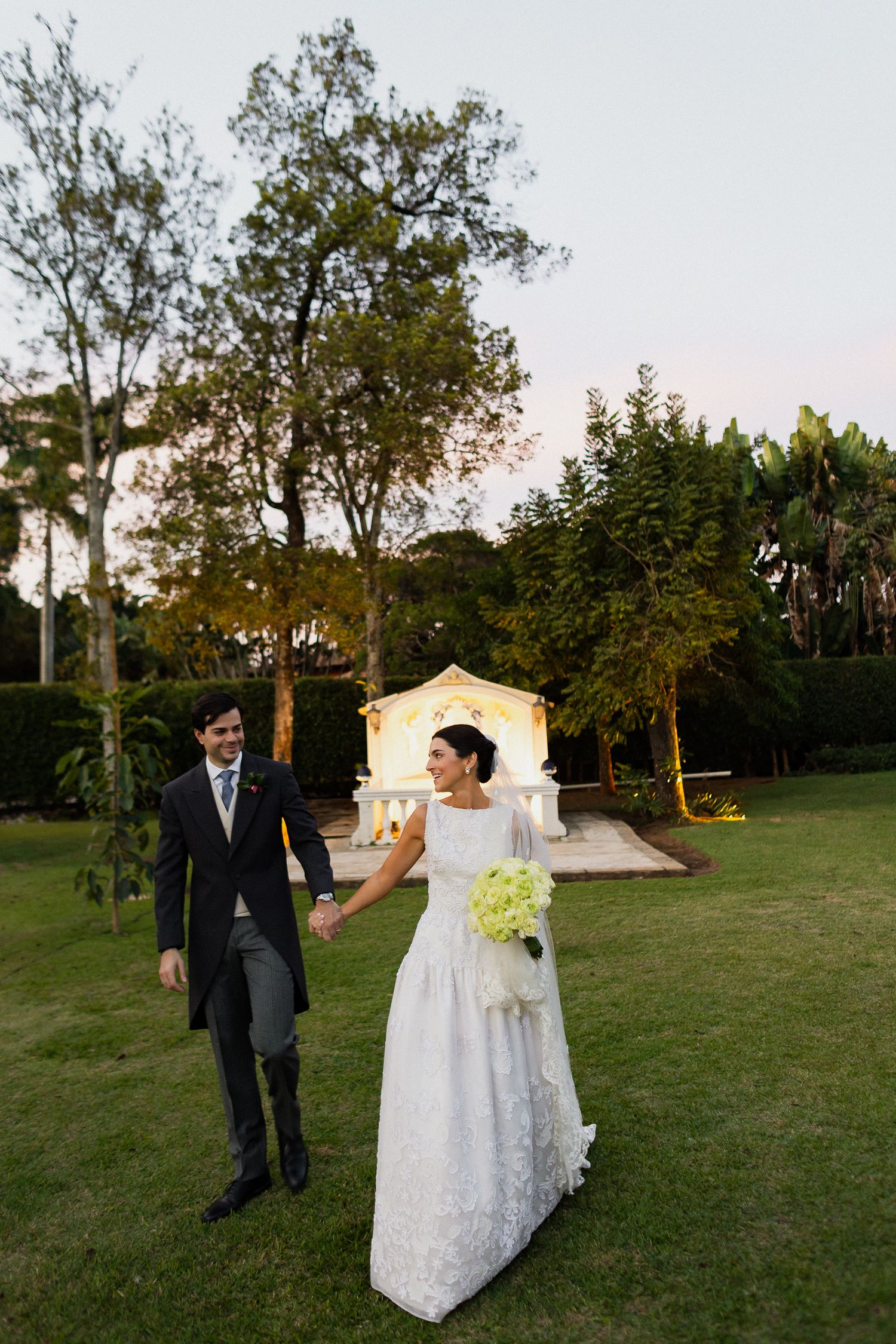 A couple walks together on a lawn with trees and a small white structure in the background during golden hour.