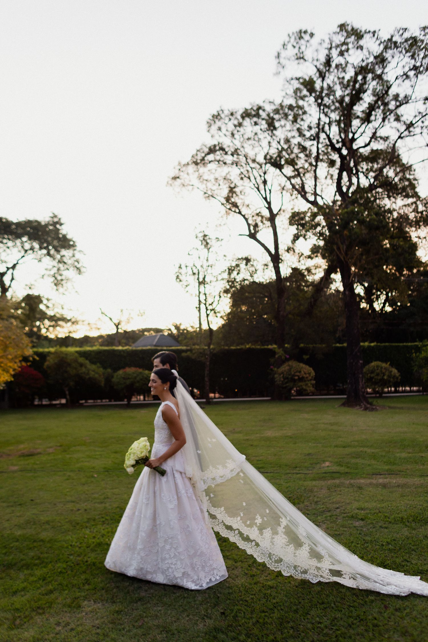 A bride in a lace wedding gown with long veil stands in a garden setting during sunset.