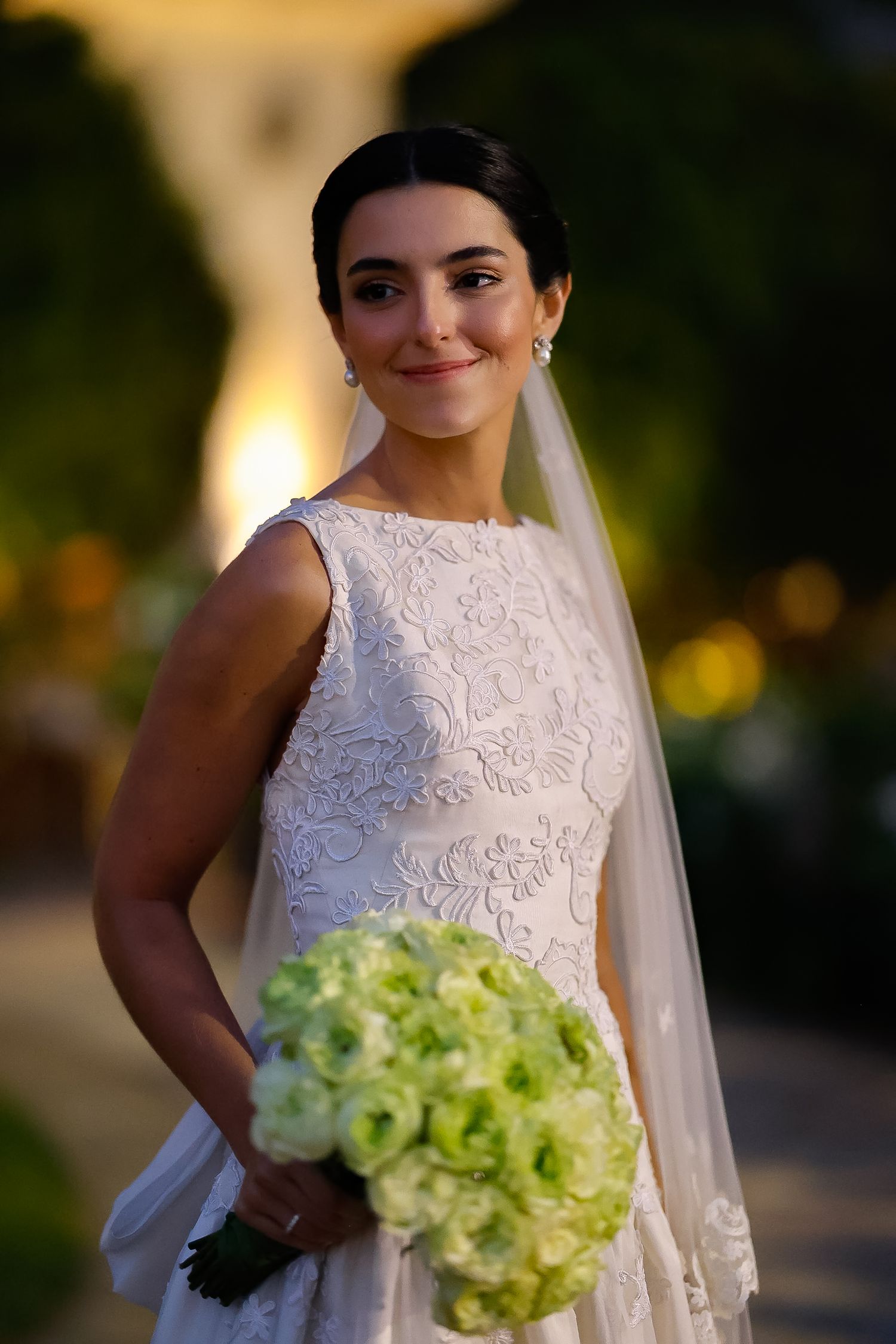 A bride in an elegant lace wedding dress holds a bouquet of green and white flowers during golden hour.
