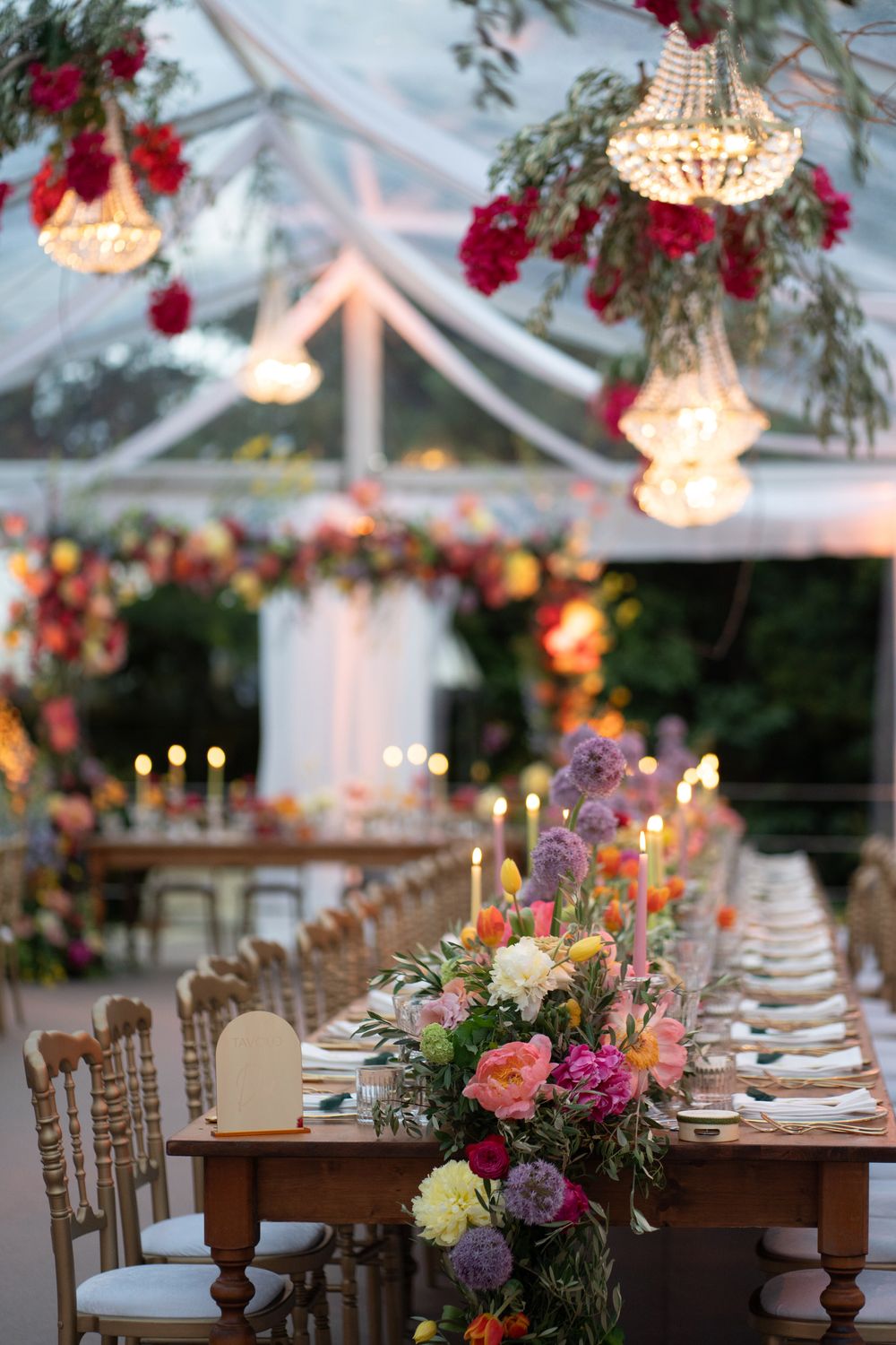 Elegant wedding reception tables set under a clear tent with crystal chandeliers and vibrant floral arrangements.