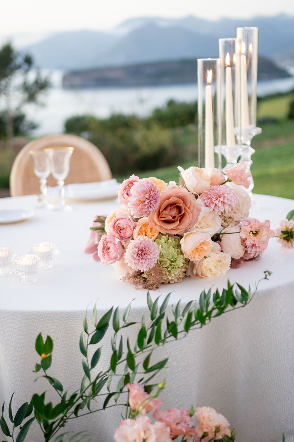 Pink floral centerpieces with roses and dahlias line a white table at an elegant outdoor wedding reception.