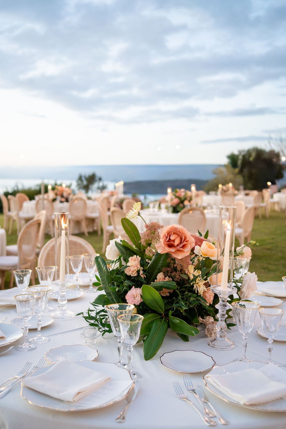Elegant outdoor wedding reception tables with peach floral centerpieces, white linens, and glassware under cloudy skies.