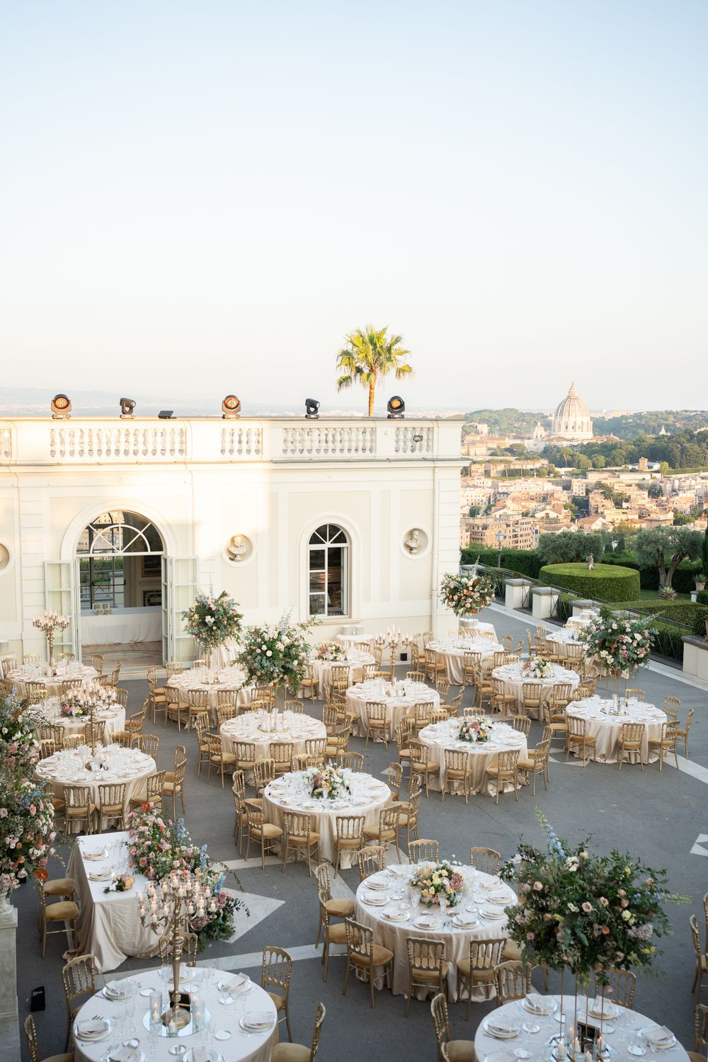 Elegant outdoor wedding reception setup with round tables and gold chairs on a rooftop terrace at sunset.