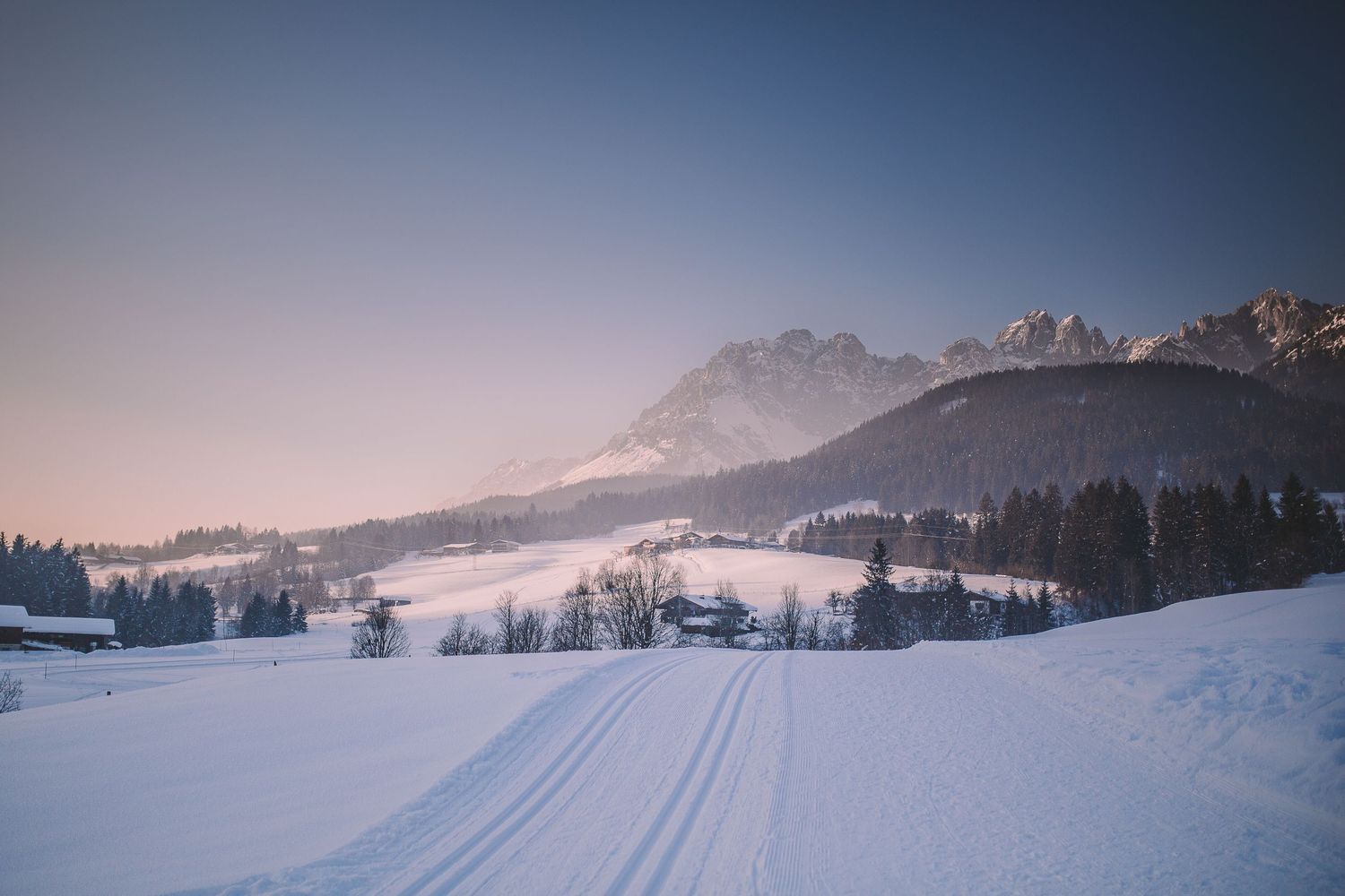 Snow-covered winter landscape with cross-country ski tracks leading toward distant mountain peaks.