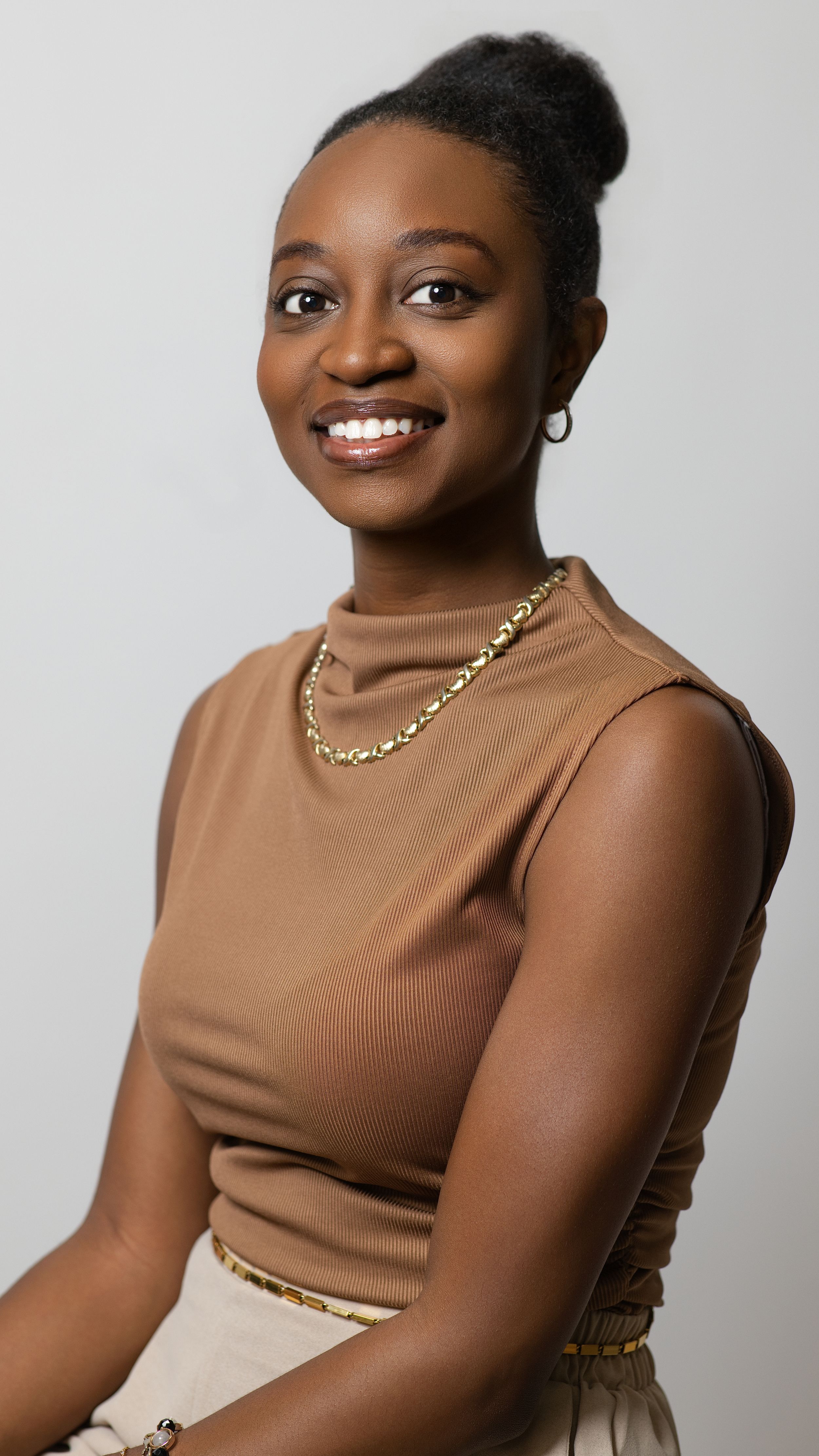 Portrait in brown sleeveless top with gold chain necklace against white backdrop.