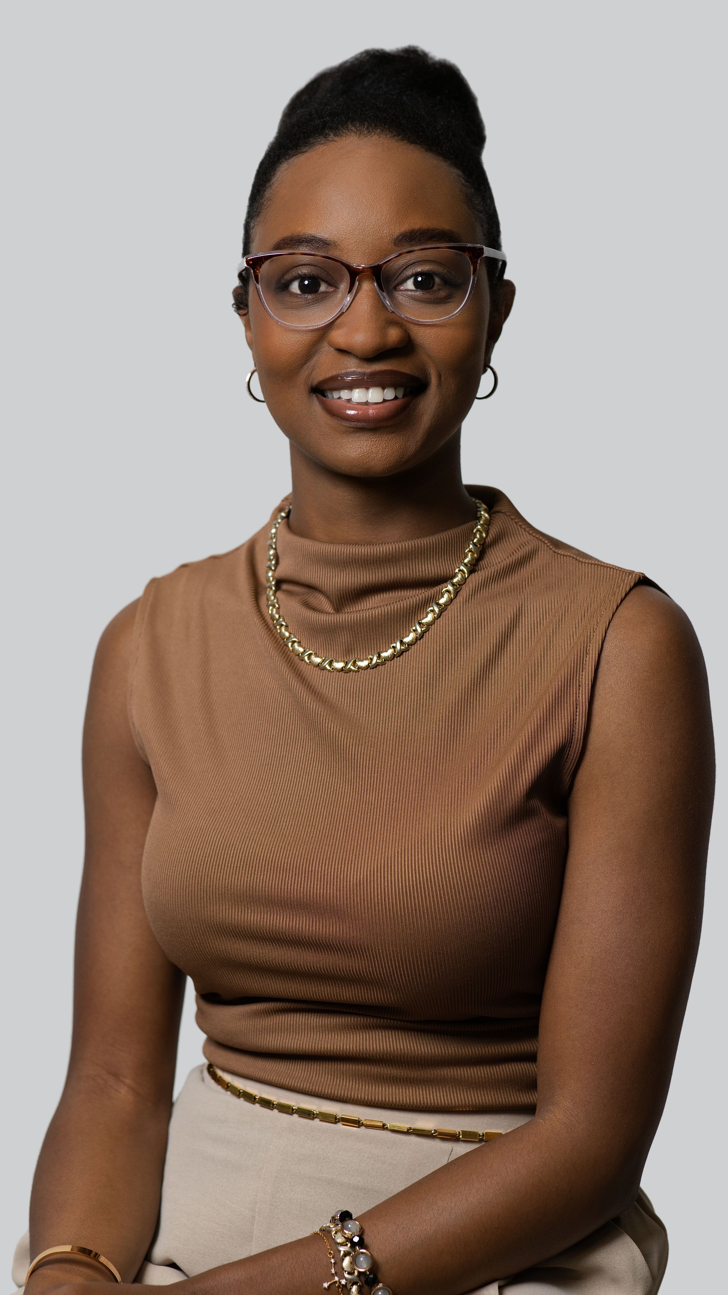 Modern portrait with brown turtleneck, glasses and gold chain necklace against grey background.