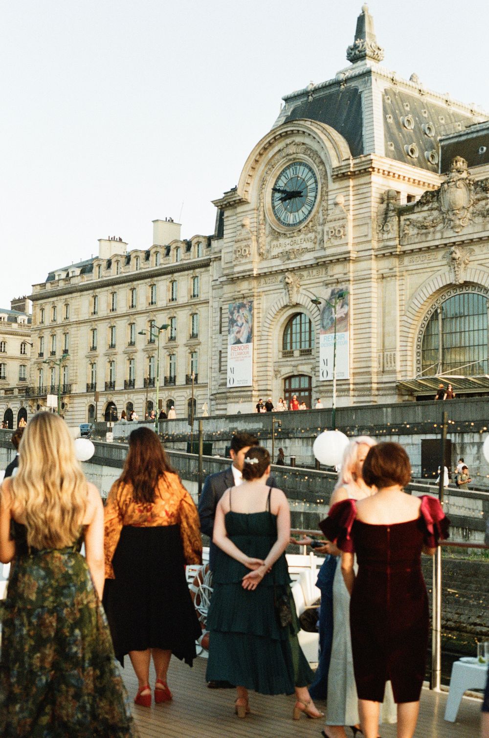 Women in elegant dresses admire the Musée d'Orsay clock facade along the Seine River in Paris, France.
