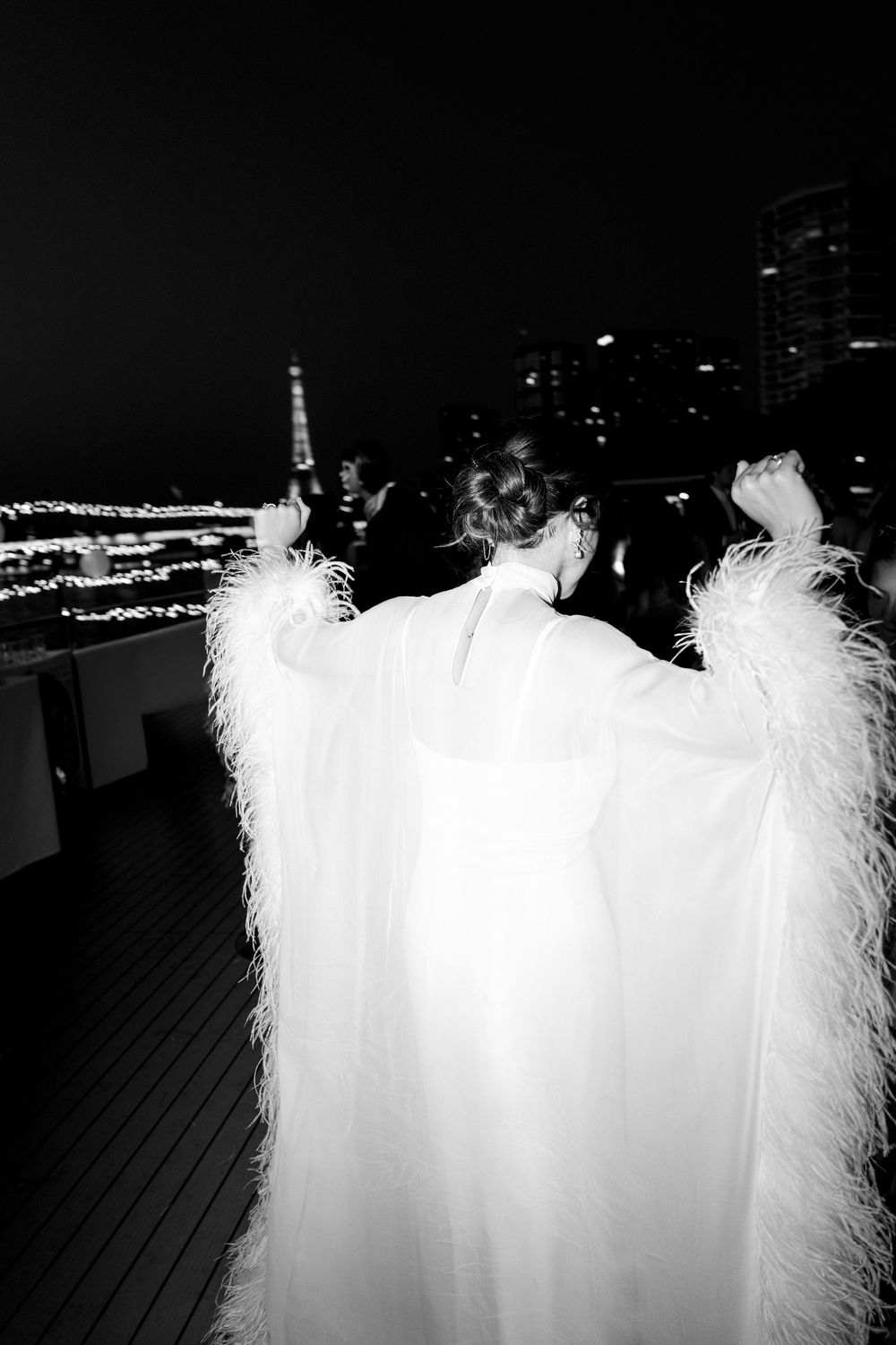 Woman in white feathered cape gown poses at night with Paris Eiffel Tower skyline in black and white photo.