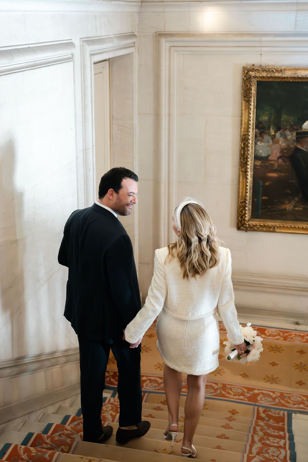 Couple in wedding attire holding hands walking through elegant hall, bride in white mini dress holding bouquet.