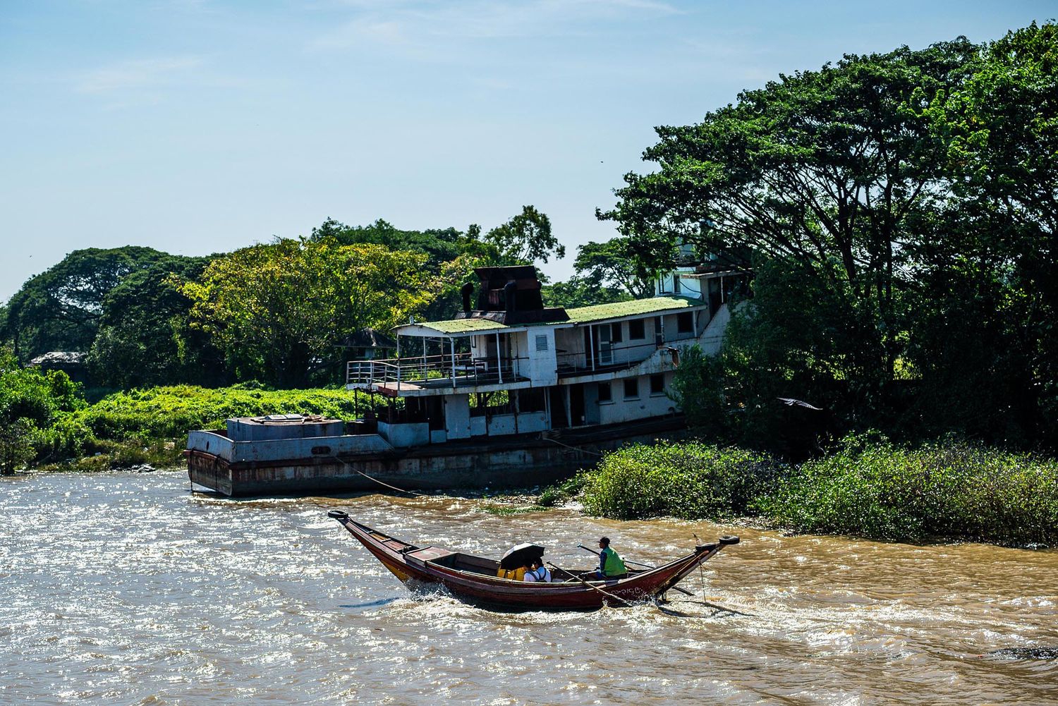 A wooden boat navigates past a riverside house surrounded by lush green trees on a sunny day in Myanmar.