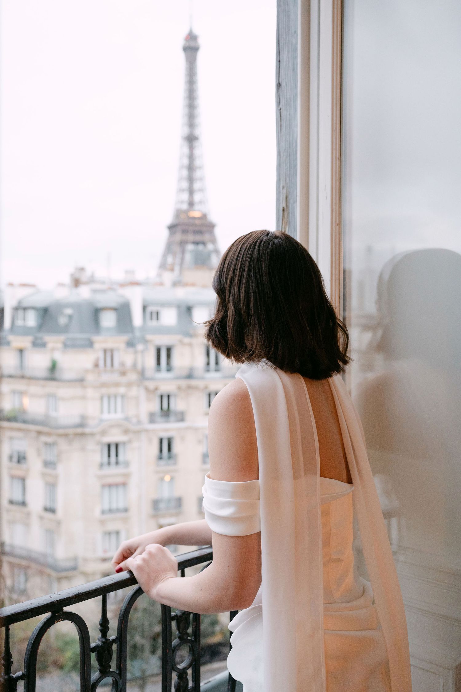 A person in a white dress gazes at the Eiffel Tower from a Parisian apartment balcony on a dreamy afternoon.