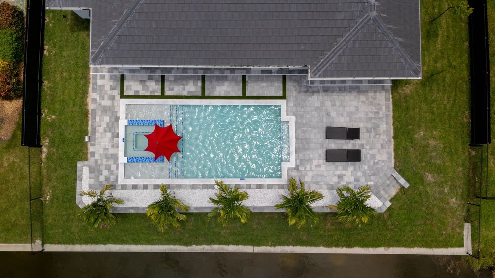 Top-down aerial view of a rectangular pool with a red umbrella and palm trees beside a modern home.
