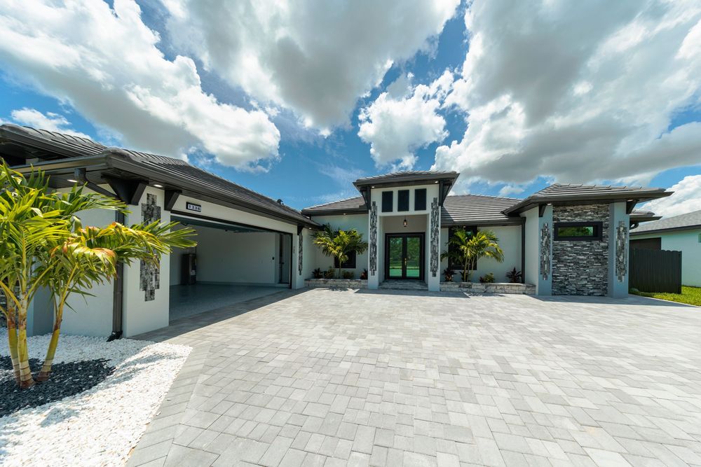 Luxury modern home exterior with paver driveway, stone accents, and lush tropical landscaping under dramatic cloudy sky.