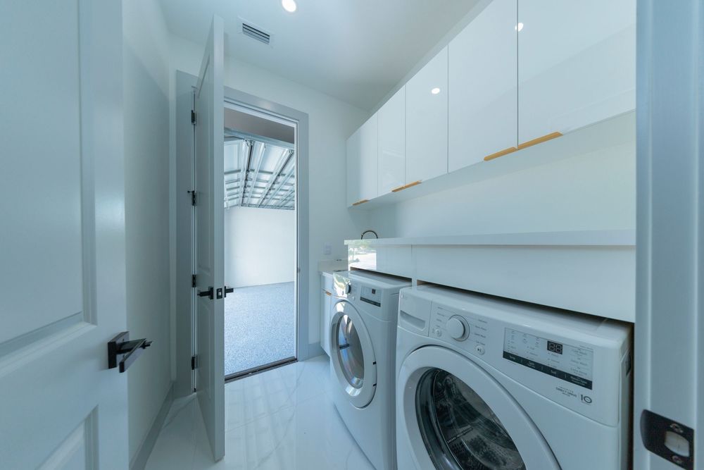 Modern white laundry room featuring front-load washer and dryer with upper cabinets and natural light.