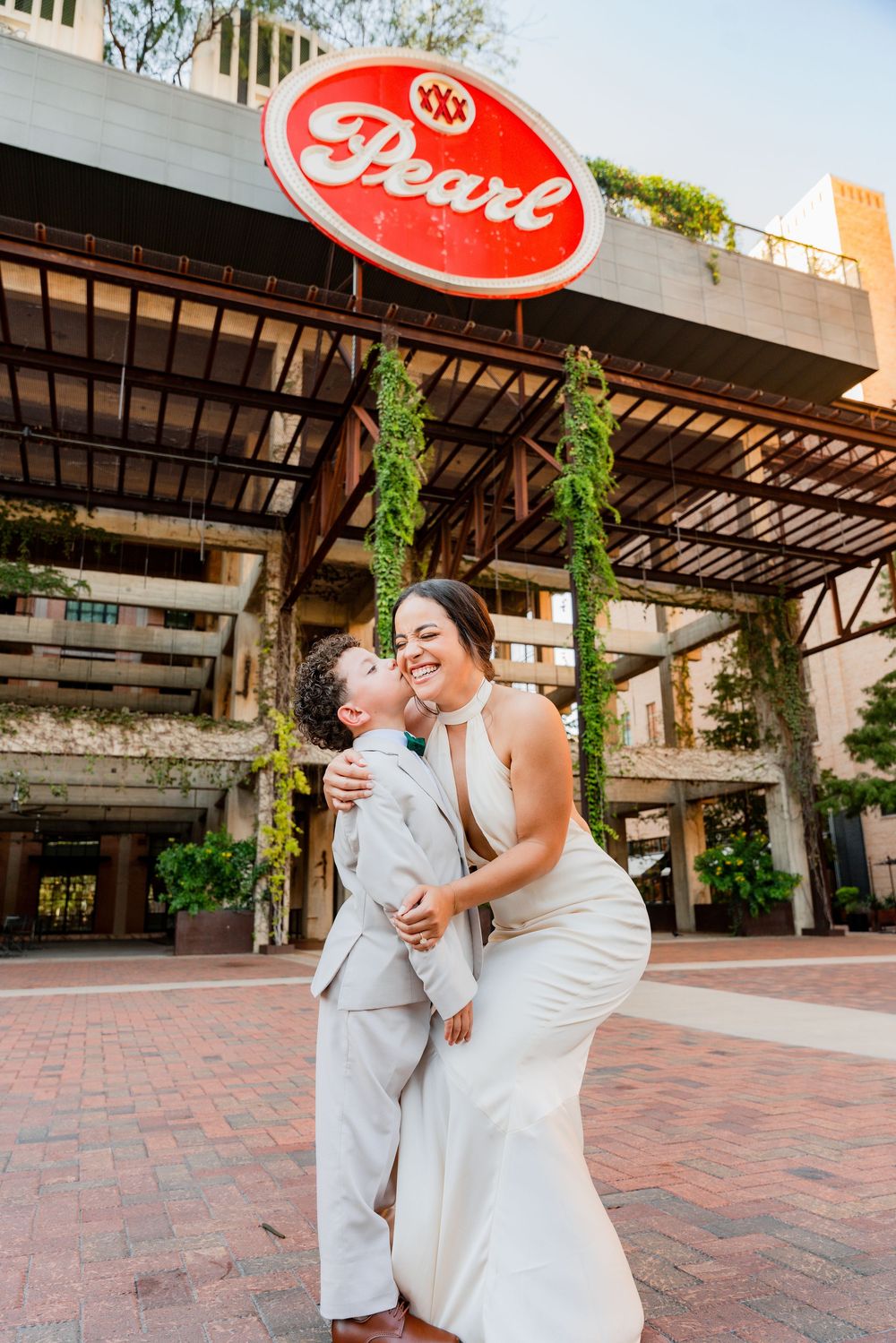 A couple in wedding attire shares a joyful moment together on a brick courtyard with wooden pergolas and greenery.