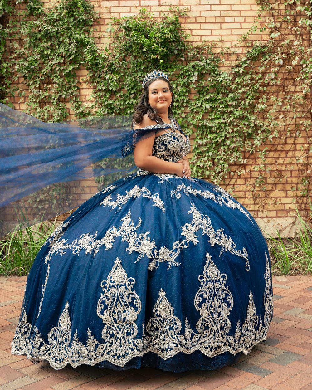 A quinceañera dress in navy blue with intricate white embroidery is showcased against a brick wall backdrop with ivy.