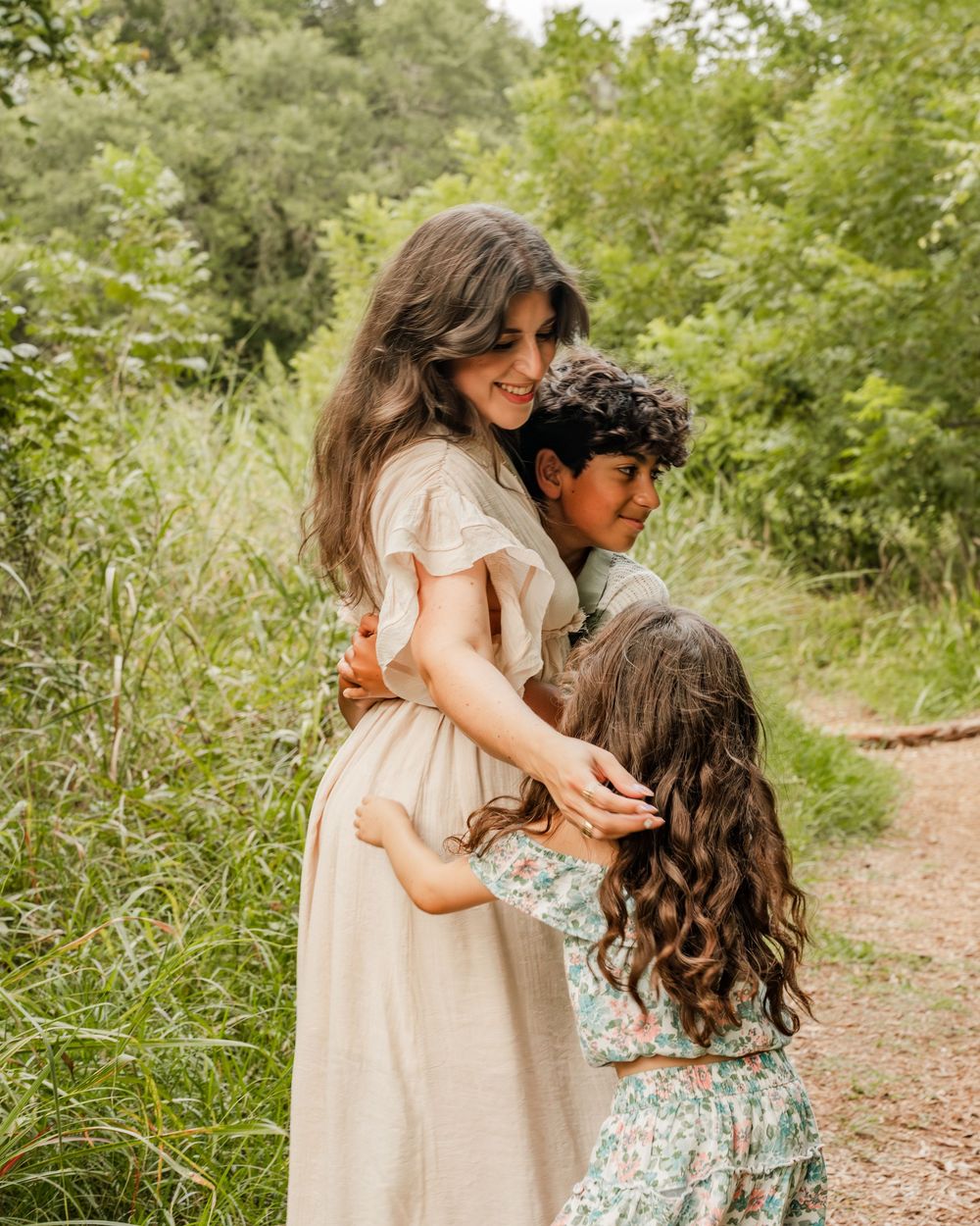 Two people share a tender embrace while walking along a sunlit nature trail surrounded by lush greenery.