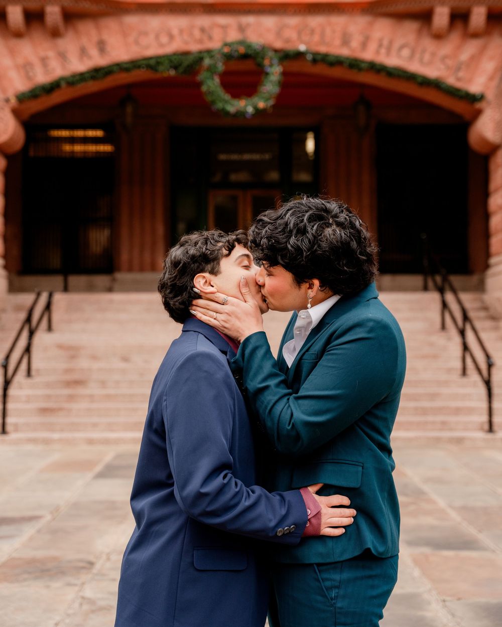 Two people in formal attire share a romantic kiss in front of a brick building with arched doorways.