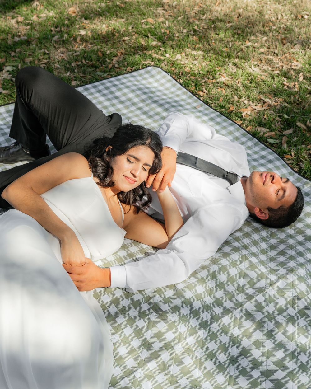 A couple lying together on a checkered picnic blanket outdoors in natural sunlight.