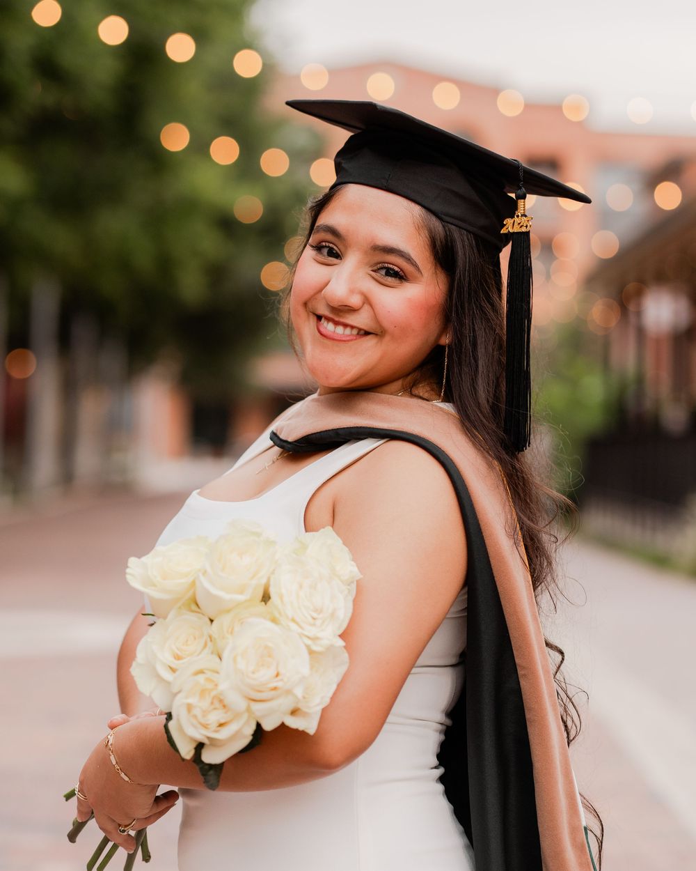 Graduate in cap and gown poses with white roses at sunset with string lights in background.