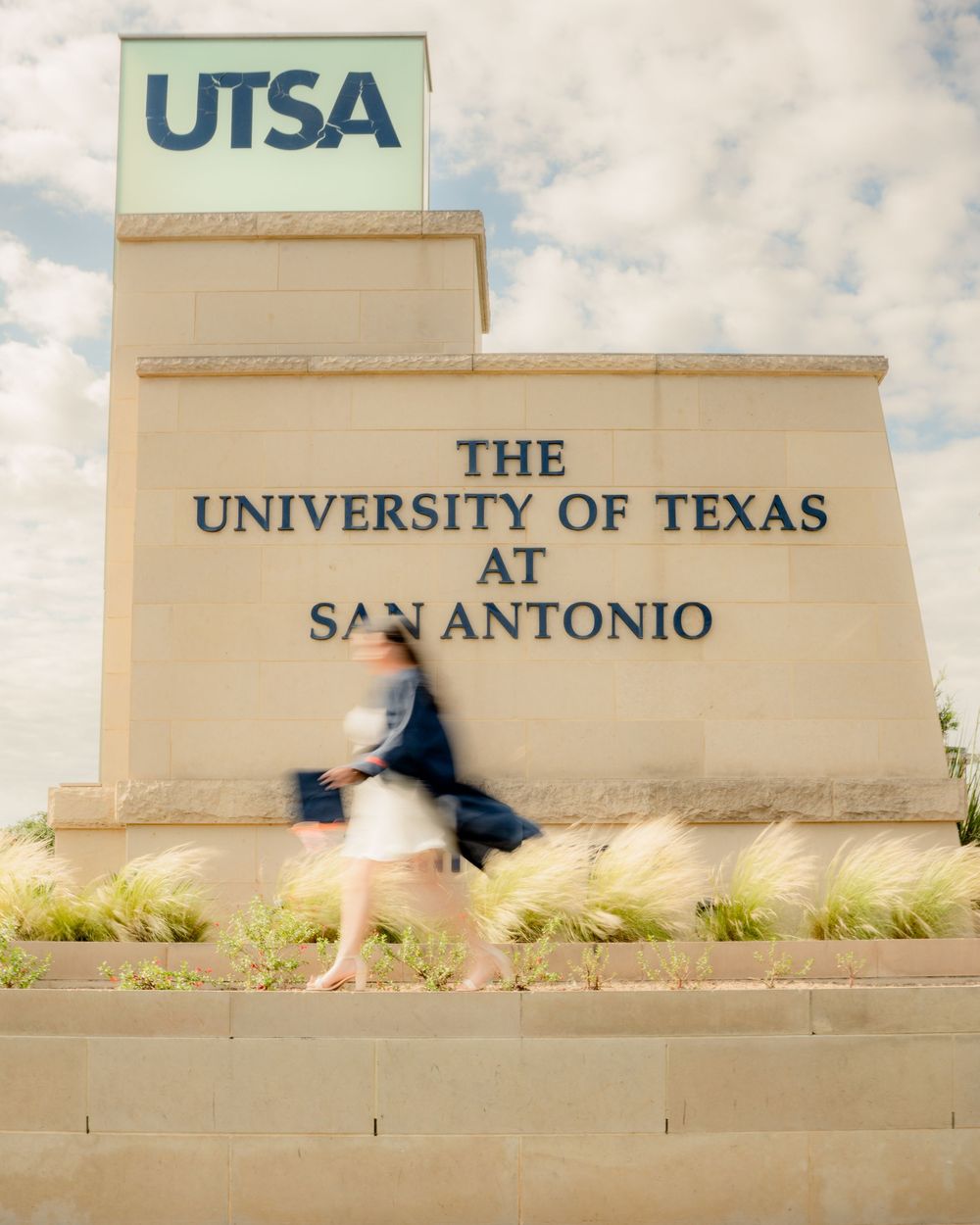 A motion-blurred figure walks past the UTSA University of Texas at San Antonio campus entrance sign.