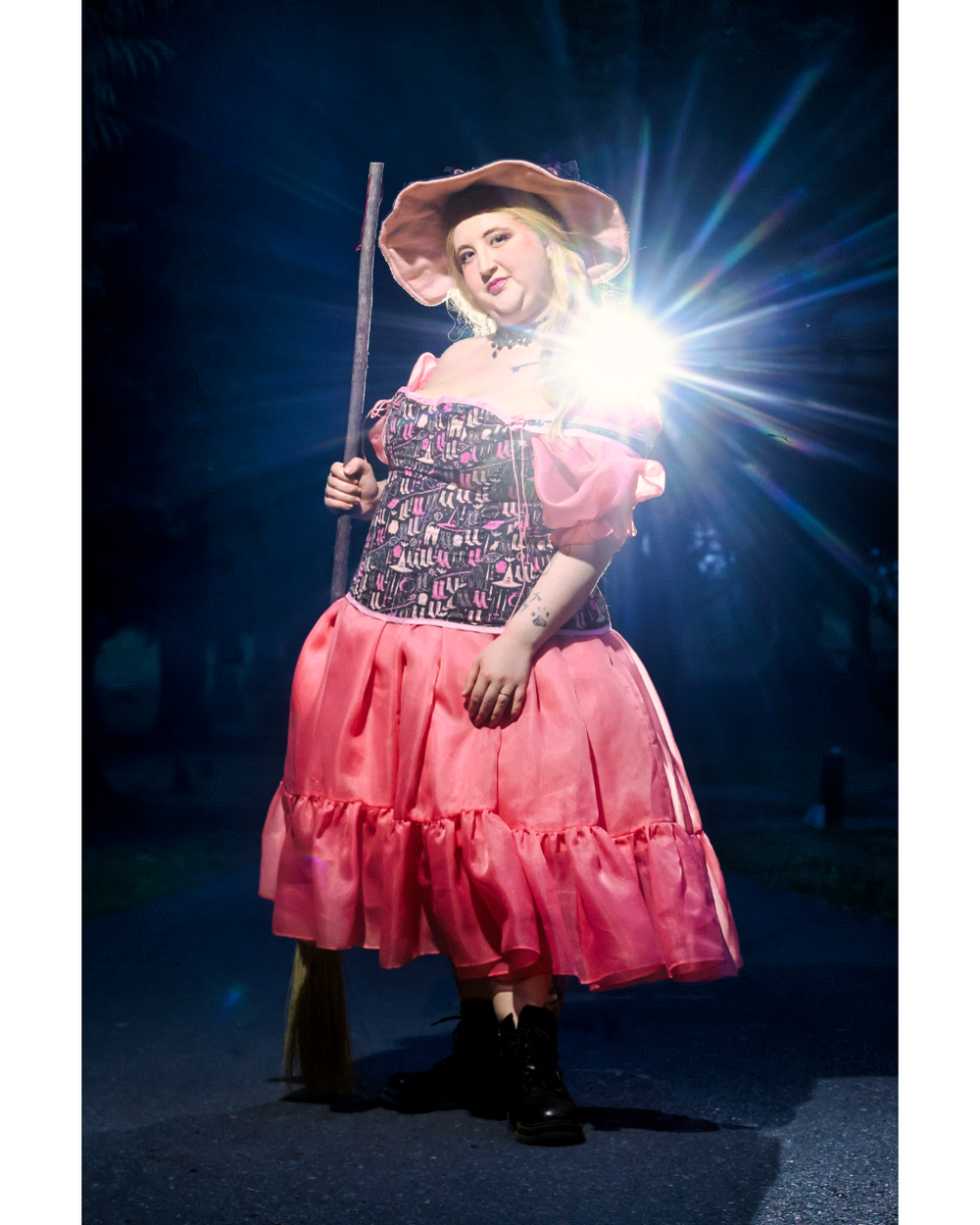 A performer in a pink ruffled dress and hat poses dramatically under a bright spotlight against a dark background.