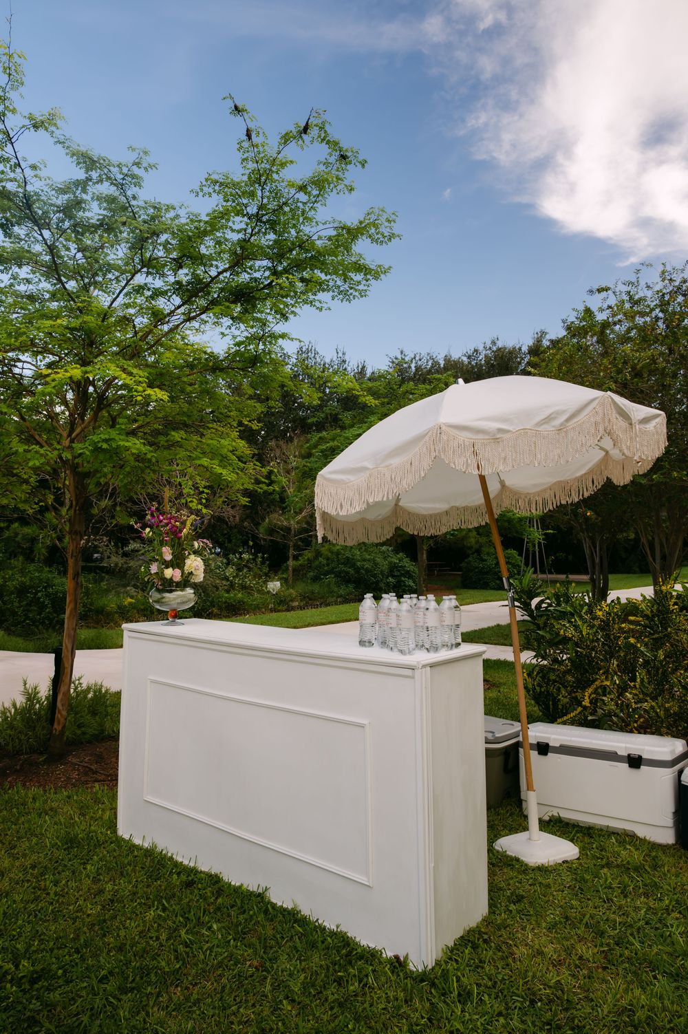 White outdoor bar setup with umbrellas against a lush green garden backdrop.