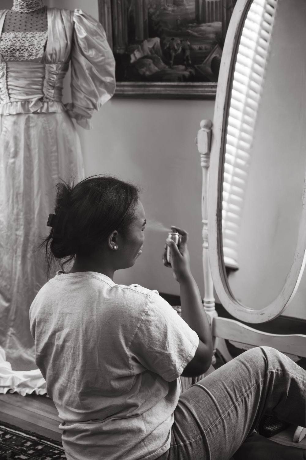 A figure sits at an ornate vanity mirror in soft window light.