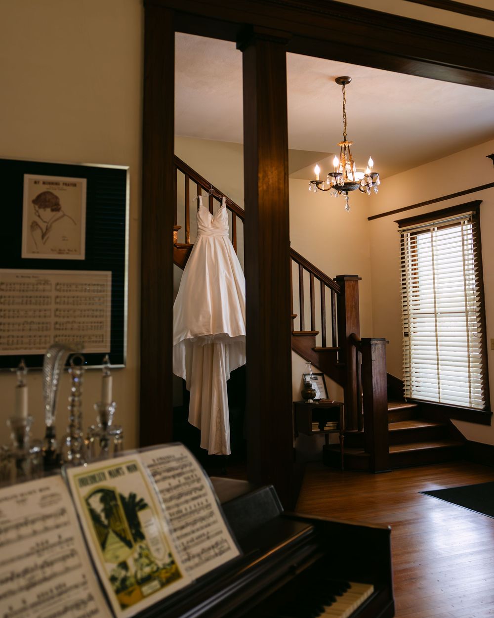 A white wedding dress hangs in a rustic interior with dark wood trim, piano, and chandelier lighting.