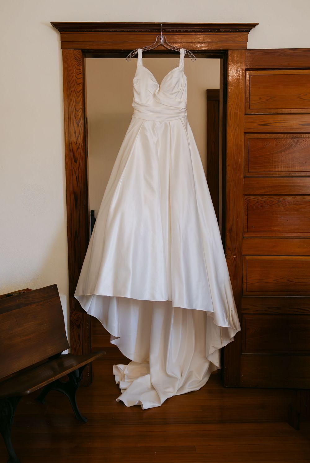 White wedding dress hanging elegantly against dark wooden furniture in a classic interior setting.