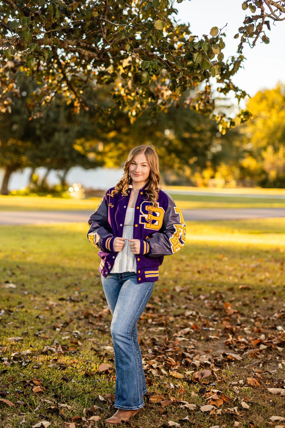 Student wearing purple and gold varsity letterman jacket poses in autumn park.