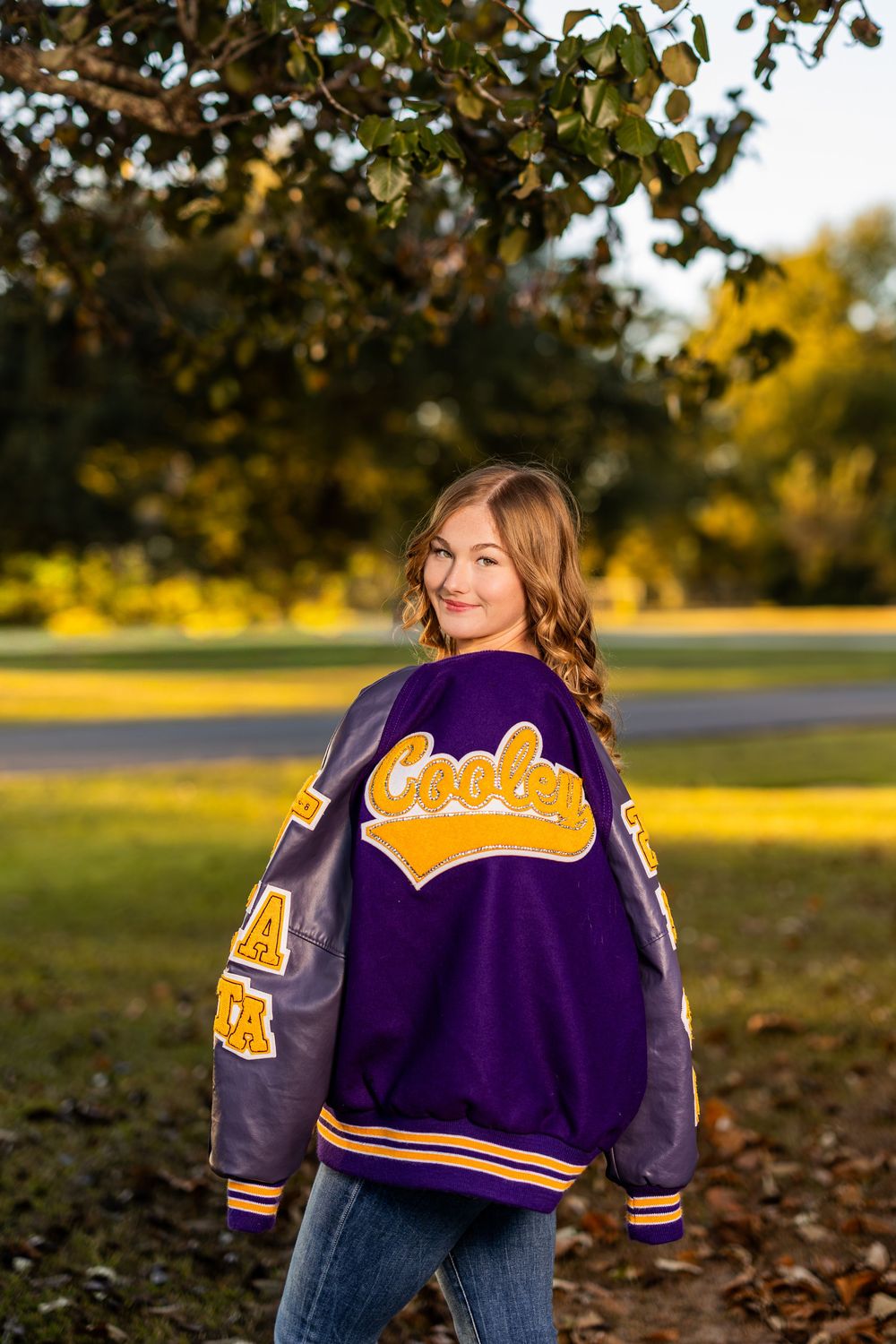 Student models purple and gold varsity jacket outdoors during golden hour.