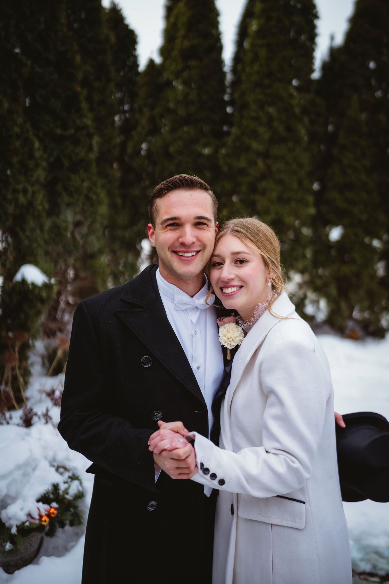 A couple in formal winter wedding attire poses together in the snow with evergreen trees in the background.