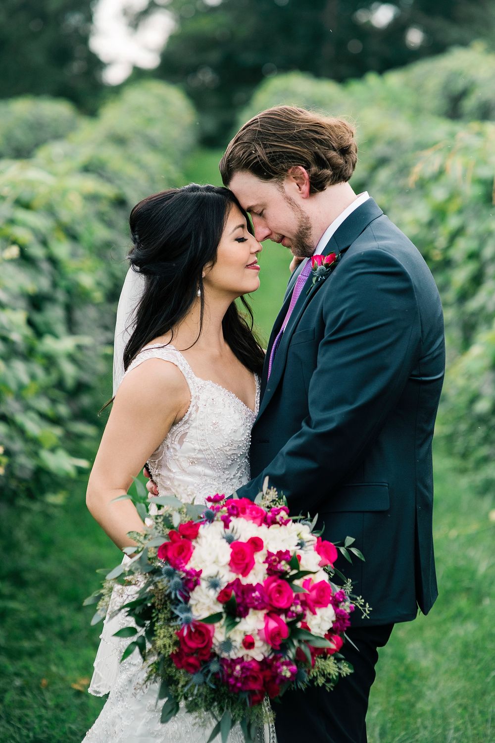 A romantic wedding couple embraces outdoors with a vibrant pink and white floral bouquet against lush greenery.