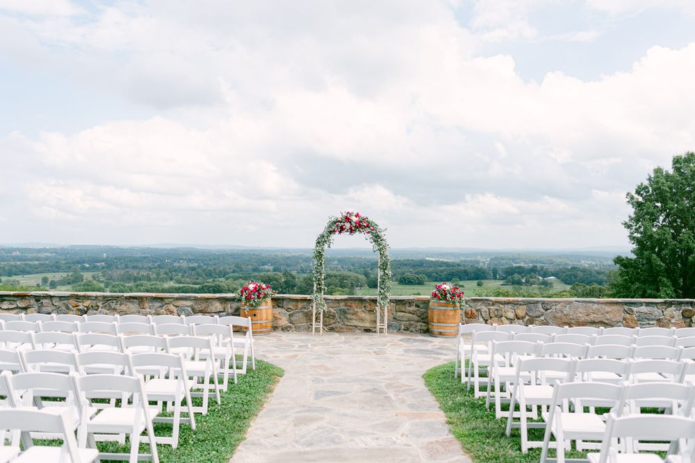 A scenic panoramic view of an outdoor wedding ceremony setup with white chairs and a floral arch overlooking rolling hills.