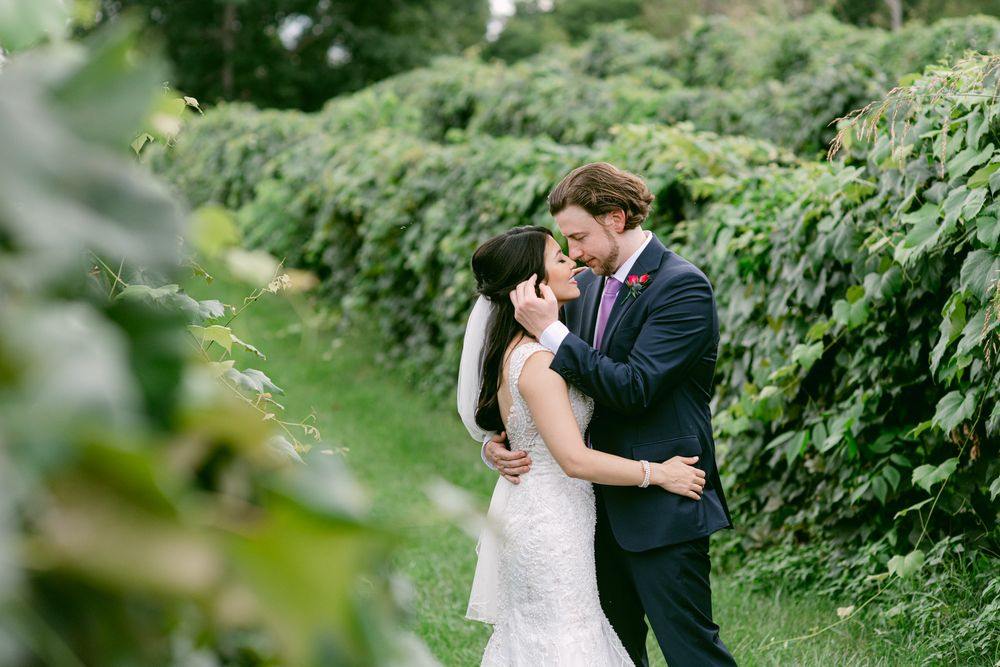 A couple in formal wedding attire shares an intimate moment in a lush green garden setting.