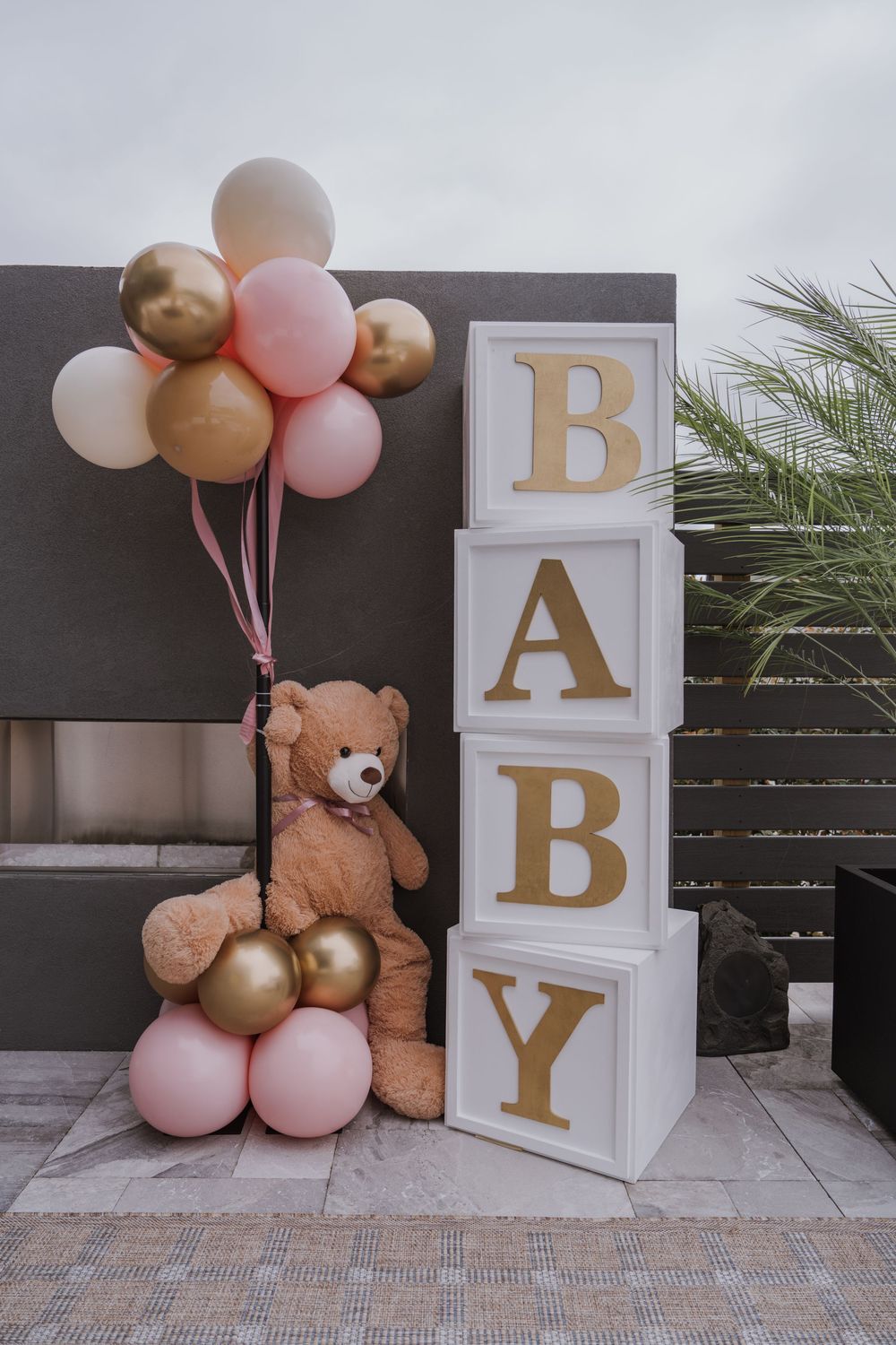 Baby shower decoration with pink and gold balloons, teddy bear, and stacked BABY blocks.