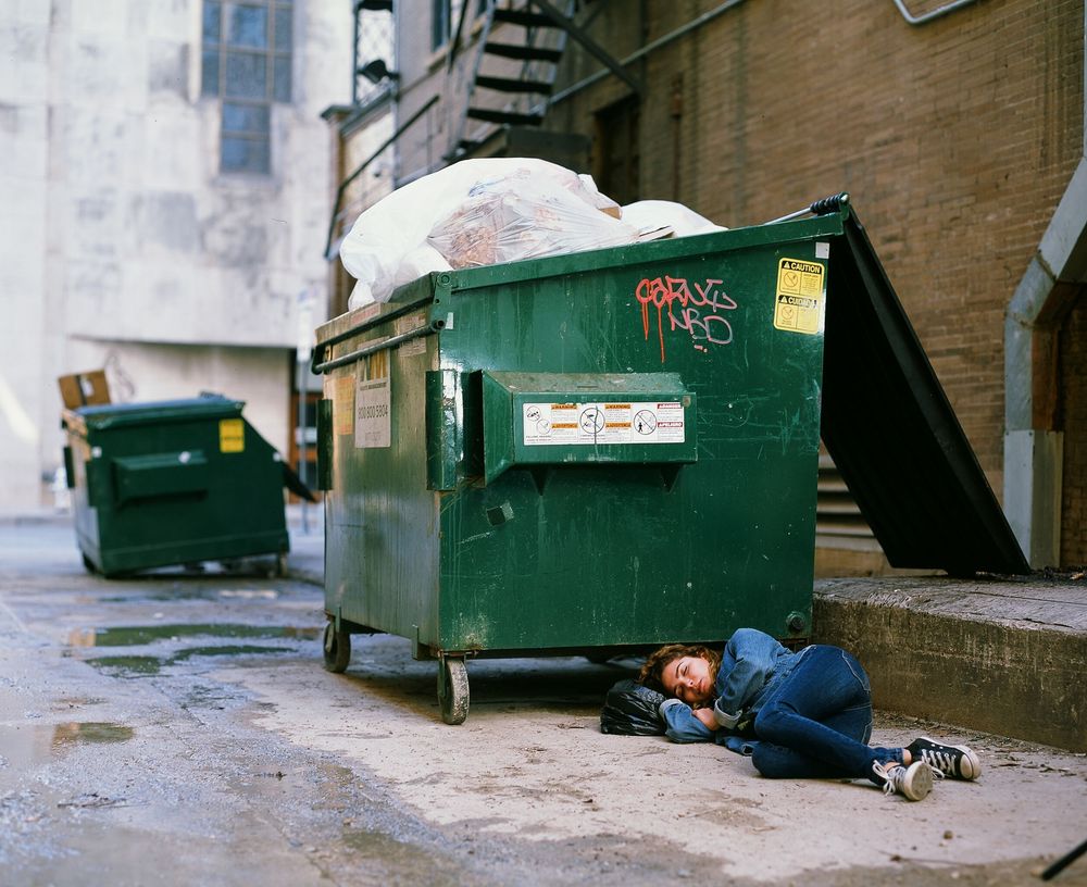Person curled up beside green dumpster in urban alleyway.