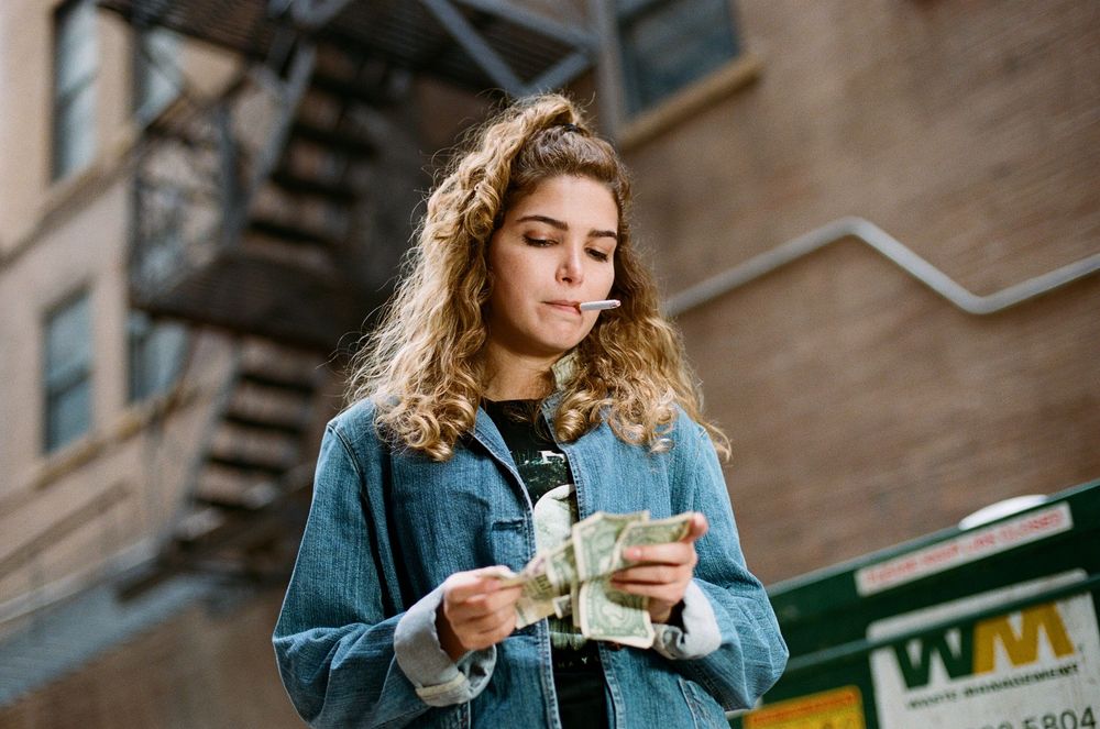 Person in denim jacket counting money outdoors near brick wall.