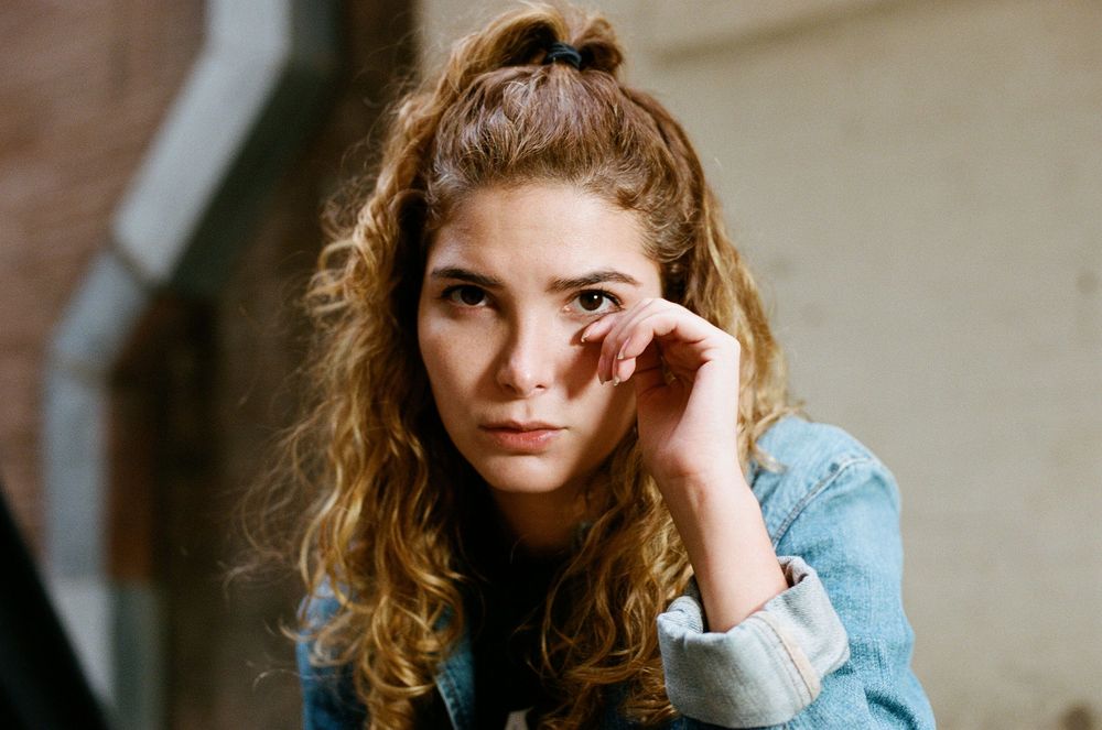 Person in denim jacket sitting thoughtfully against beige wall.