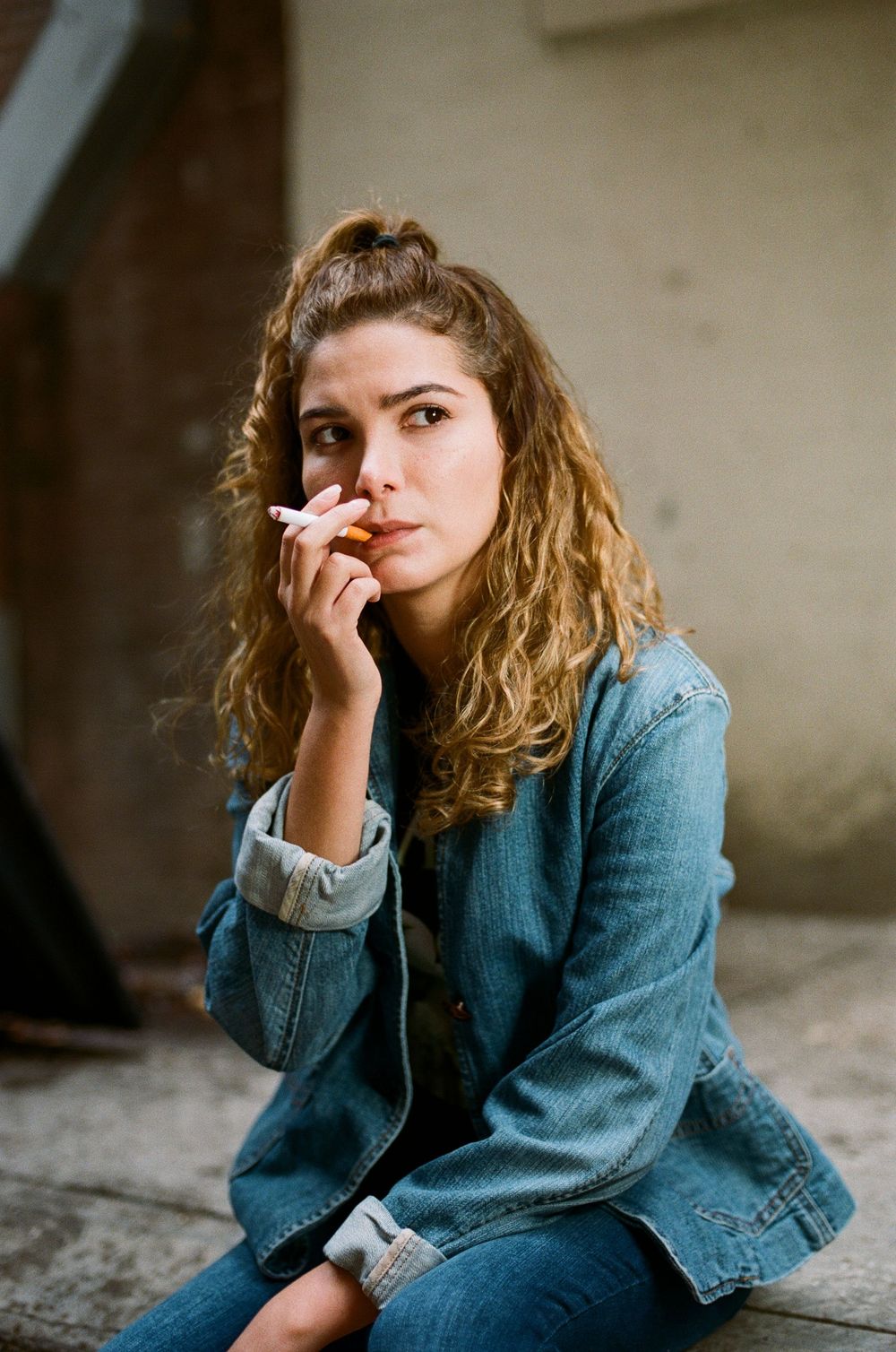 Person in denim jacket sitting pensively against wall with curly hair.