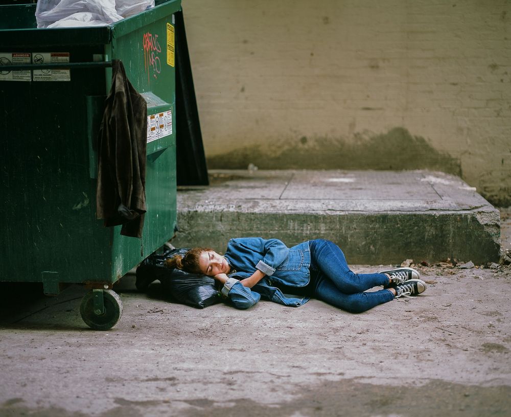 Person in denim lying on ground next to green dumpster in urban setting.