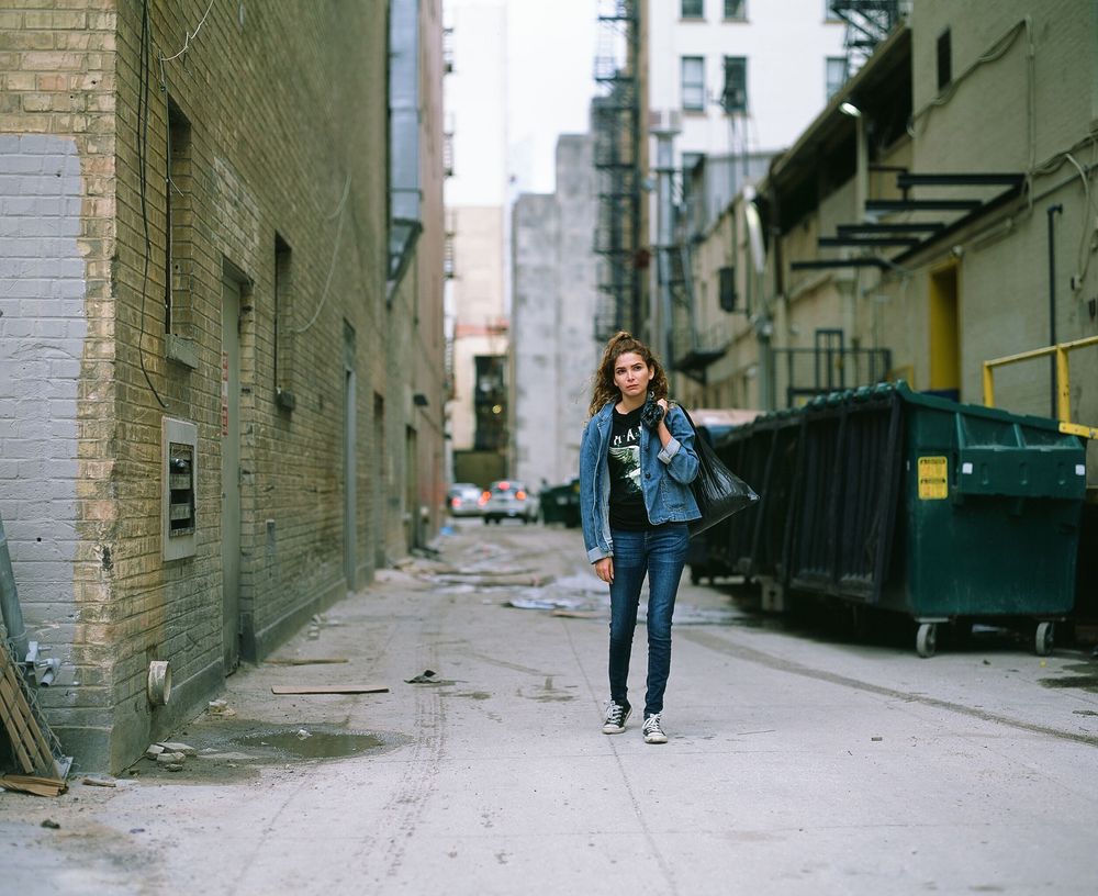 Figure walking down urban alley with brick walls and dumpsters.